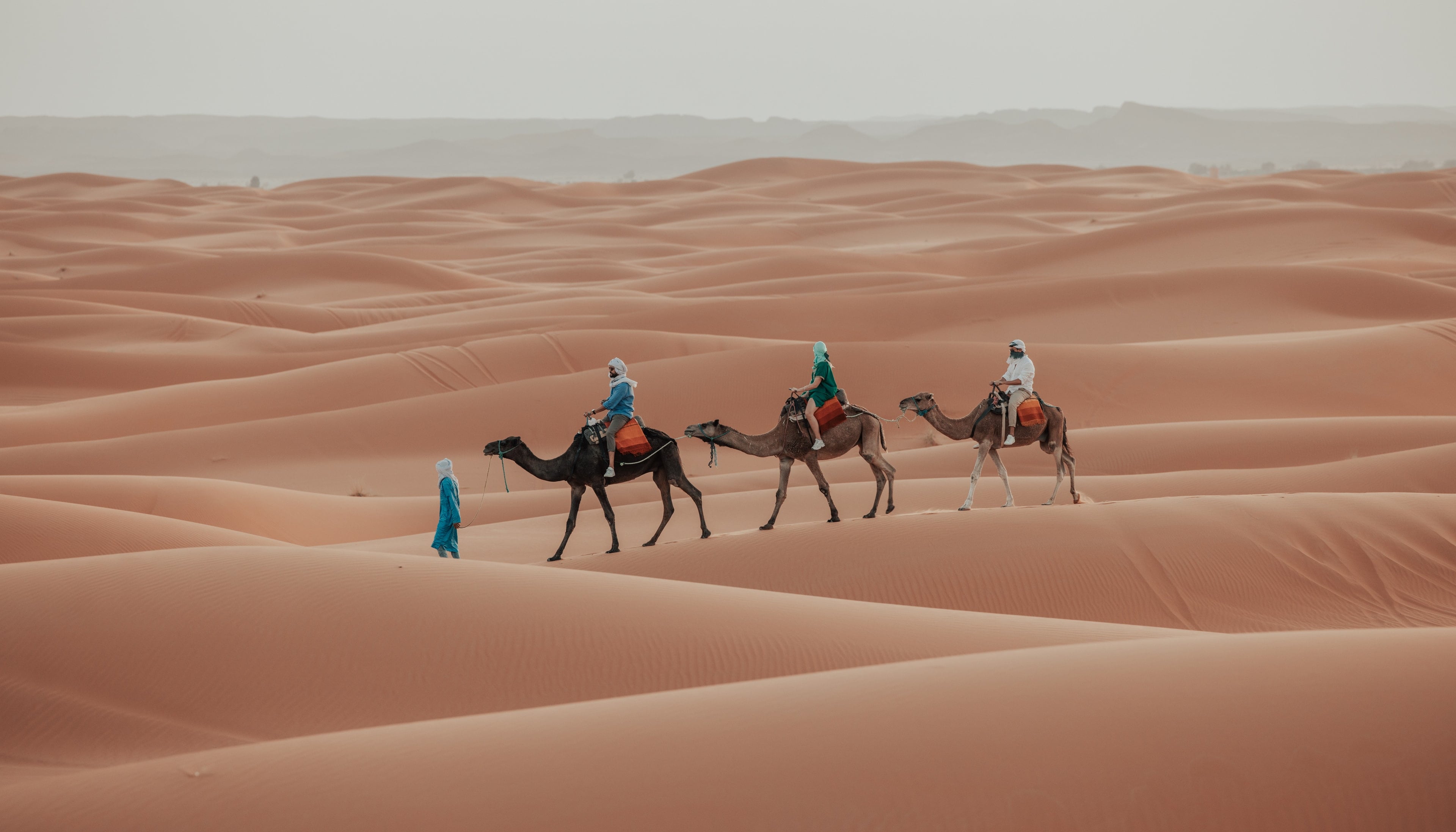 Three riders on camels crossing pale dunes in a soft haze