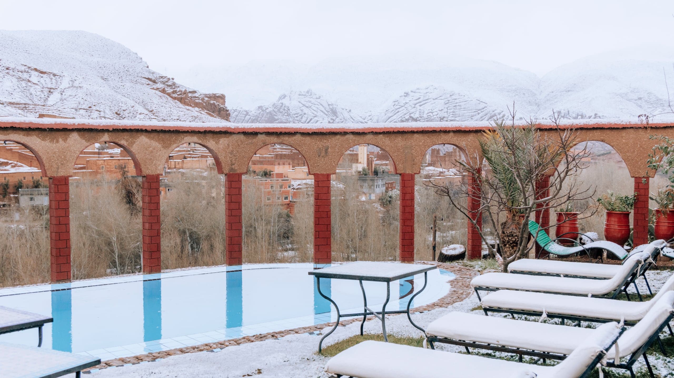 A hotel pool with arches and loungers dusted with snow, mountains behind