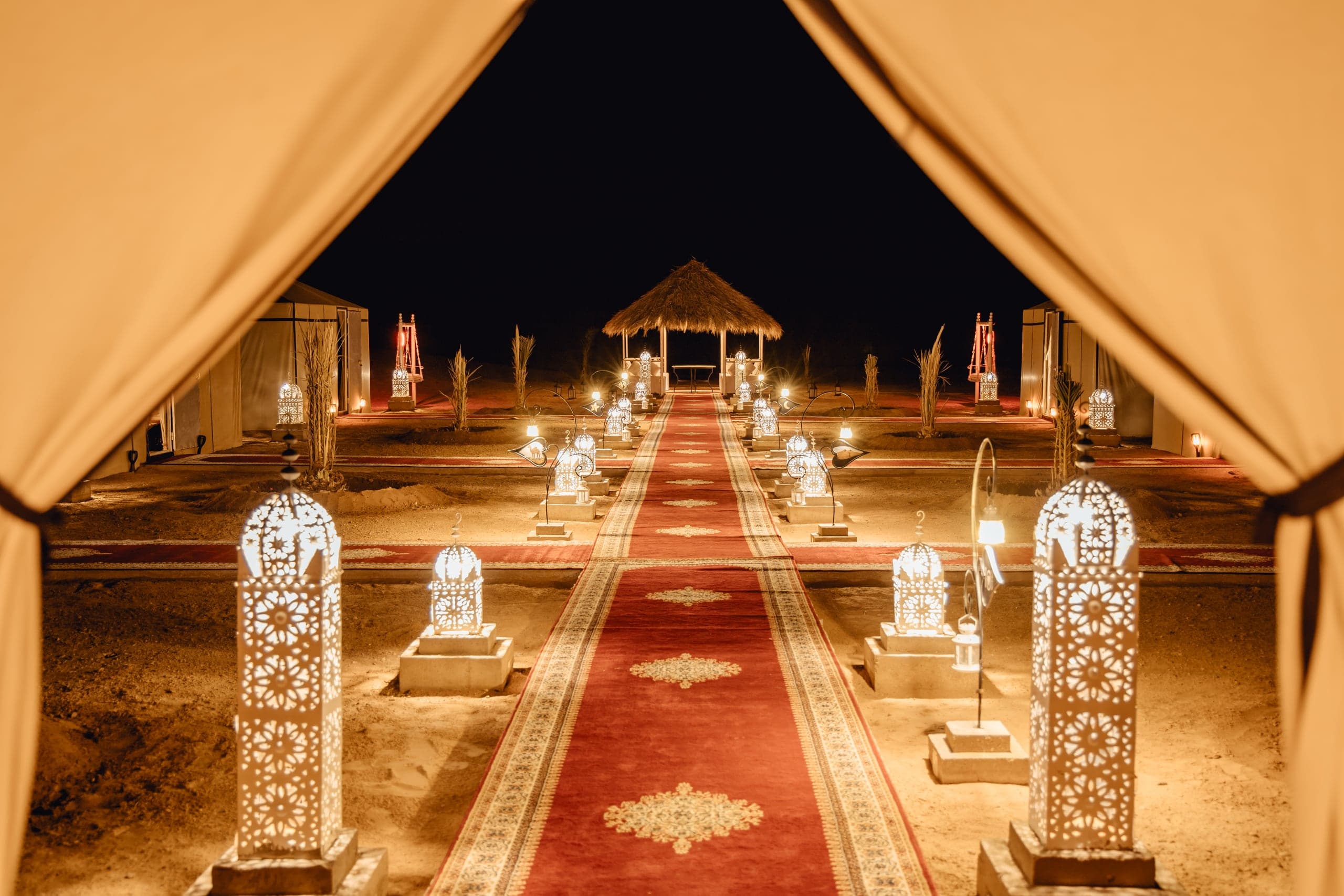 Lanterns and a red carpet leading through a desert camp at night, seen from a tent