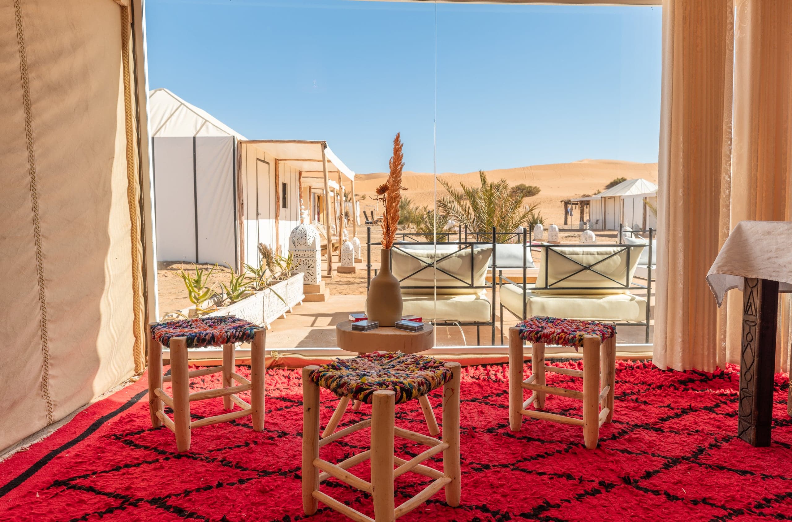 Inside a camp tent: a red Berber rug and stools facing the dunes