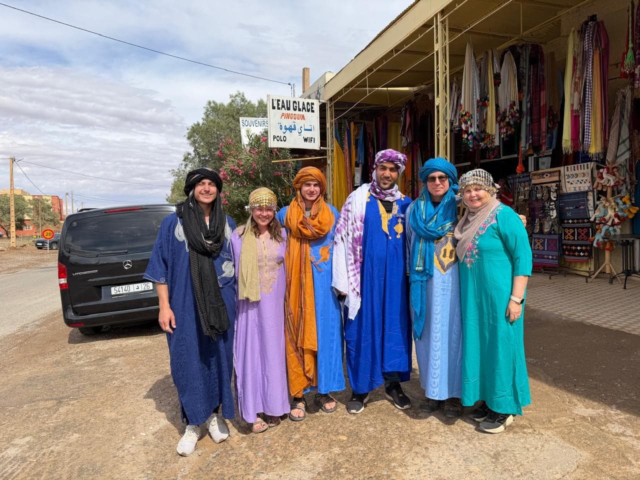 Guests and their driver in Berber robes and turbans outside a roadside shop, the van parked beside them