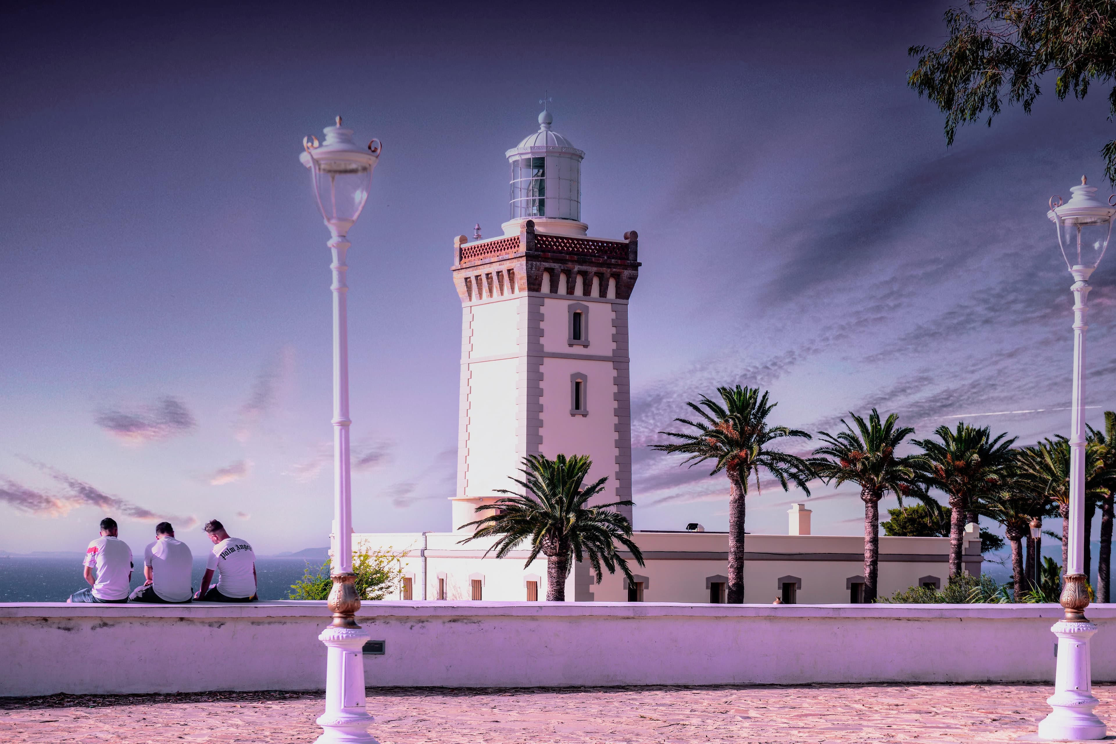 The Cap Spartel lighthouse near Tangier at dusk, with palms and three people on a wall